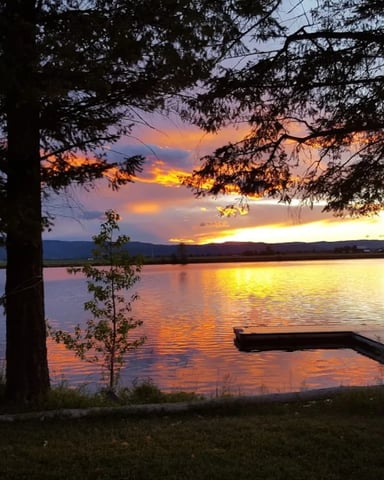 Sunset over a calm lake with a dock and hammock, framed by pine trees